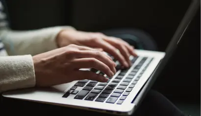 Close-up of a person typing on a laptop. The hands are placed on the keyboard, and the screen is partially visible. The person is wearing a light-colored long-sleeve garment. The background is slightly blurred.