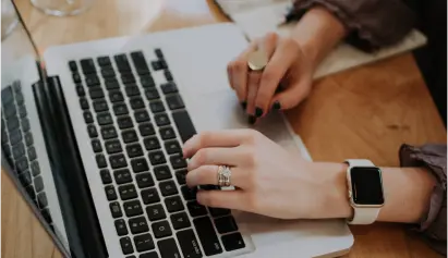 A person is typing on a laptop keyboard while wearing a smartwatch on their left wrist. They have rings on several fingers of their left hand. A notebook with a pen is partially visible in the background. The surface they are working on appears to be wooden.