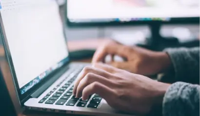 Person in a grey sweater typing on a laptop keyboard, with a blurred screen visible in the background. Focus is on the hands and keyboard.